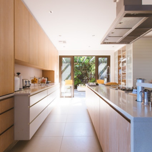A long galley kitchen with light wood upper cabinets, white lower drawers and a large set of storage shelves with opaque glass doors off to one side.