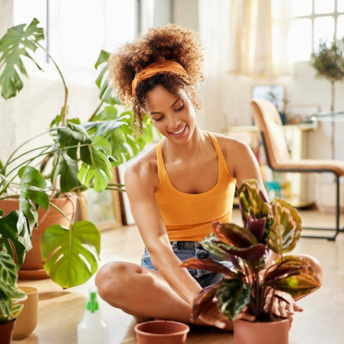 a young Black woman, with her hair tied back, sitting cross-legged on the floor of her apartment with her plants