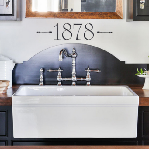 A deep white porcelain farmhouse kitchen sink with beveled silver faucets. The sink is surrounded by dark wood countertops and black cabinetry with vintage silver drawer pulls.