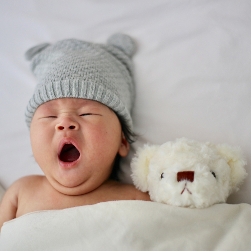 Newborn baby tucked into bed with a stuffed toy and yawning