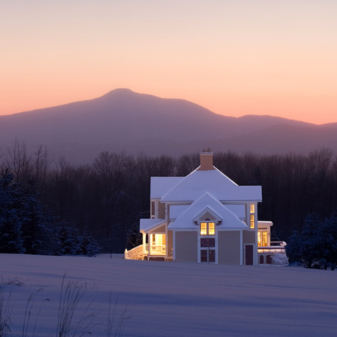 A snow-covered house in the countryside against a mountainous backdrop during sunset.