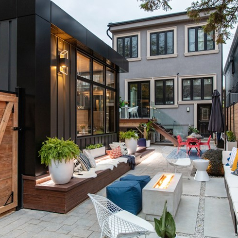 A cool backyard lounge area with a large white outdoor couch, two teal square poufs, a built-in wooden bench seat with sheepskin throws, cement paving stones, tall wooden fence with black hardware, large potted plants, and a black backyard laneway house as featured on Backyard Builds on HGTV Canada