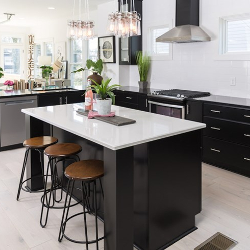 Chic modern kitchen with light wood floors, a large centre island with three barstools and a white countertop, black cabinets, black counters, black and stainless-steel appliances, and two groups of glass pendant lights as featured on Master of Flip on HGTV Canada