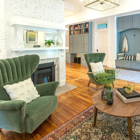 Beautiful living room featuring stained hardwood floors, two green velvet wing backed chairs, a red woven rug, a round wooden coffee table, a white brick fireplace, a blue coat nook, and white walls as featured on Property Brothers on HGTV Canada