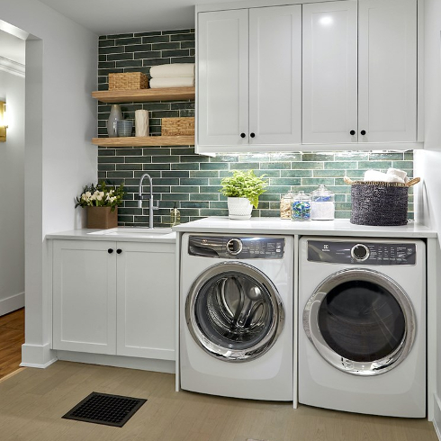 Pretty laundry room featuring a white wash and dryer installed on the floor with a countertop and soaking sink, green tile backsplash, white storage cupboards, and wood open shelving stacked with towels, baskets and group of vases as featured on Farmhouse Facelift on HGTV Canada