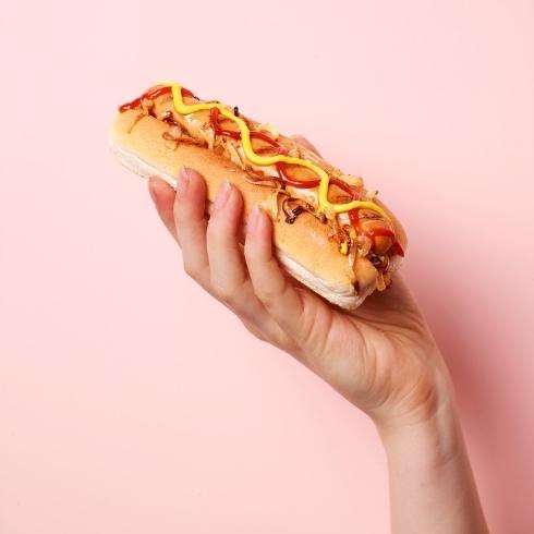 A person holding up a hot dog with ketchup and mustard in front of a pink background.