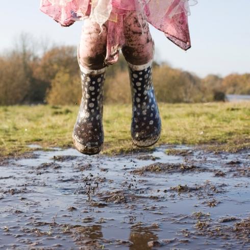 A kid jumping in a mud puddle with a pink jacket and muddy green rainboots.