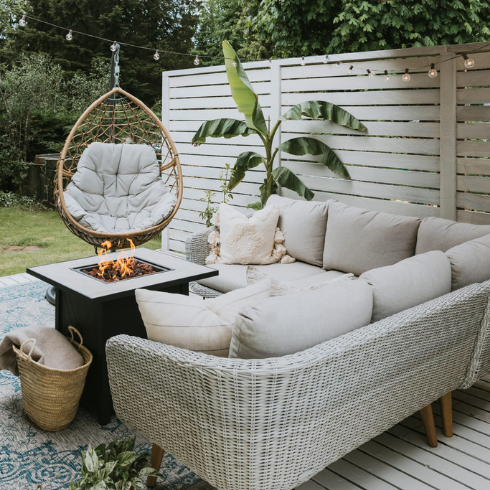 A back deck with a privacy paint, all painted off-white. Furnished with a white-beige rattan outdoor sectional sofa with cream upholstery, a black firepit, and a rattan hanging chair.