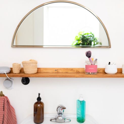 Small bathroom with wood shelf above sink and gold half crescent mirror