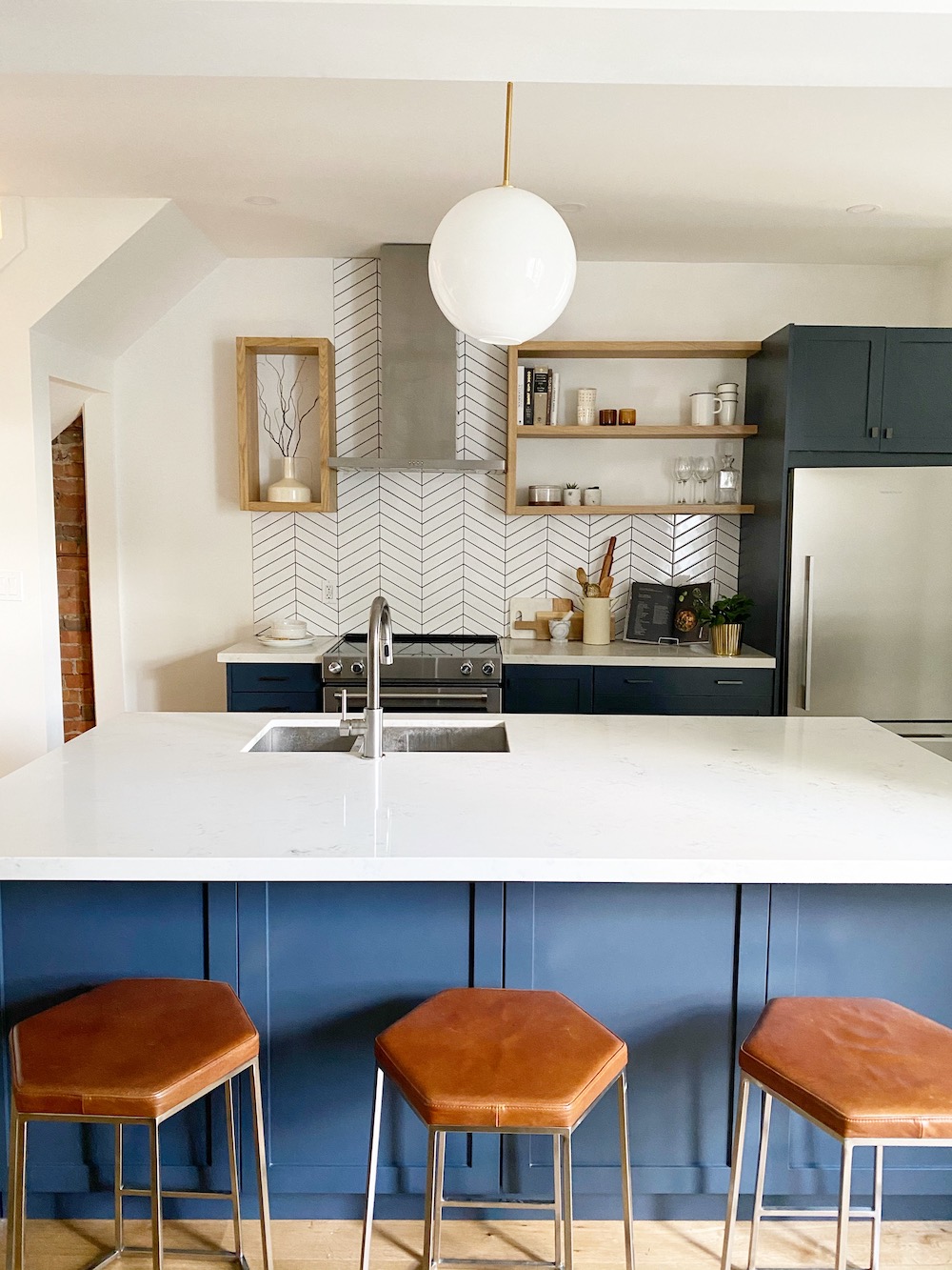 Modern kitchen designed and styled by The Property Stylist Inc. with large blue island with a white quartz countertop and undermounted kitchen sink, dark blue cabinets, white glass globe pendant light, and white herringbone tile backsplash