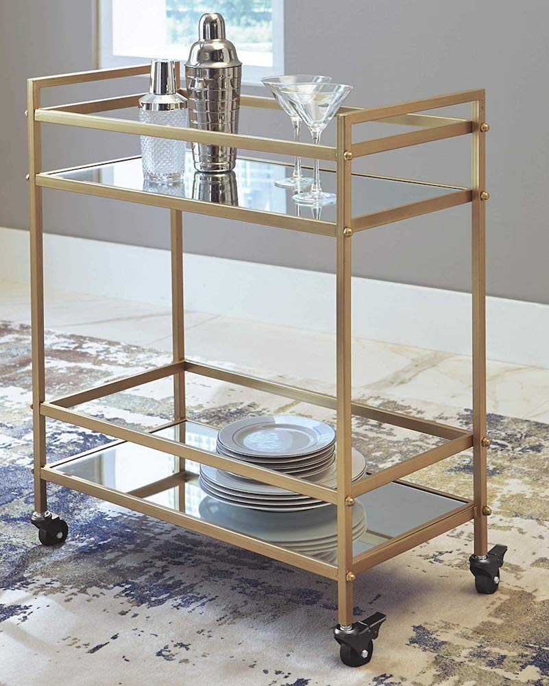 Gold car cart with caster wheels and two mirrored shelves holding a stack of white plates, two cocktail shakers and two martini glasses