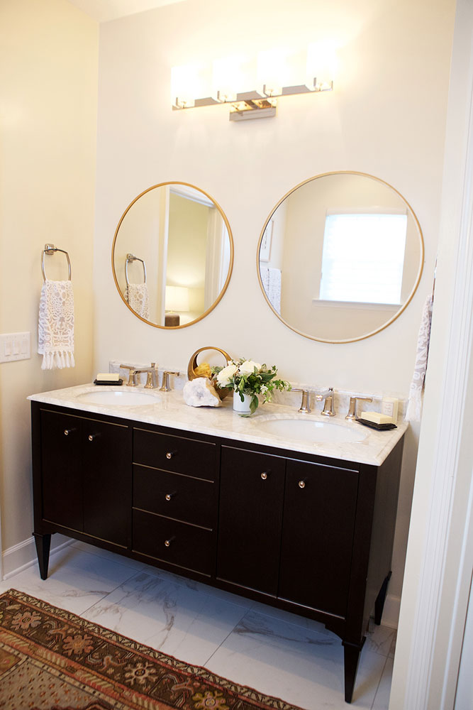 Modern bathroom with a double sink black vanity, large marble floor tiles, a woven rug and two round gold mirrors