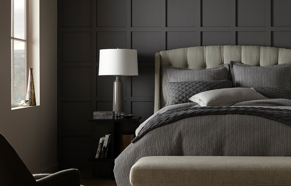 Moody modern bedroom with upholstered bed layered with grey bedding, a bedside table with a grey and white lamp, and walls painted in BEHR Mined Coal PPU18-18 and Silver Marlin N360-2