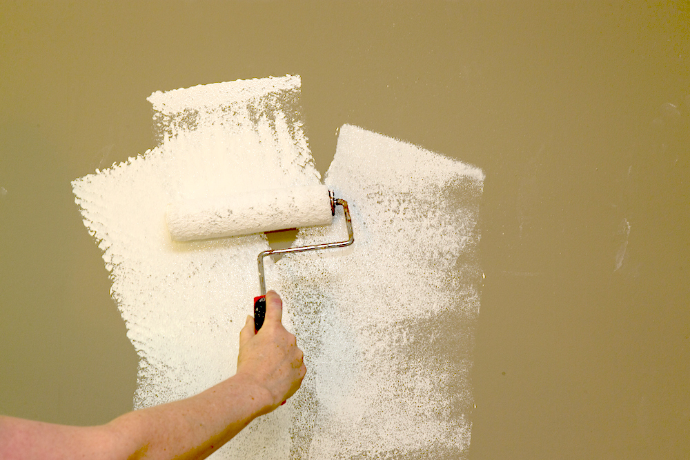 A hand paints white primer with a paint roller onto dark painted wall