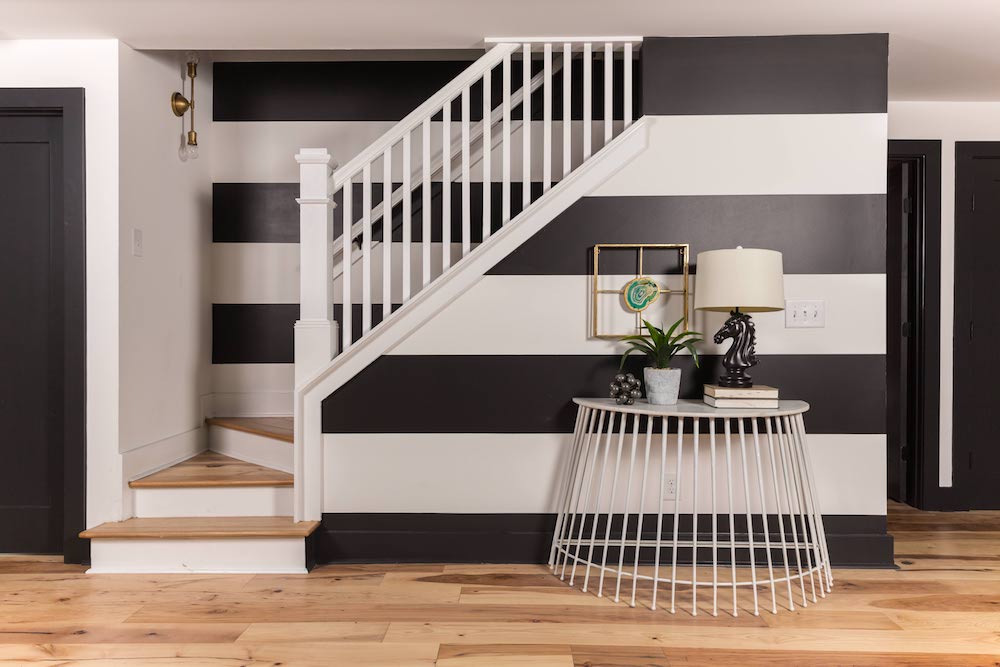 Striking staircase painted in wide black and white stripes with a white hall table set at the bottom with a horsehead lamp, a small stack of books, a potted plant, and a framed agate slice as featured on Masters of Flip on HGTV
