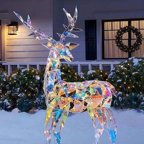 72-inch Christmas sparkle modern angular-iridescent-reindeer buck standing in a snowy front yard in front of a classic white craftsman house with lights in the hedge and wreaths on the front door and window