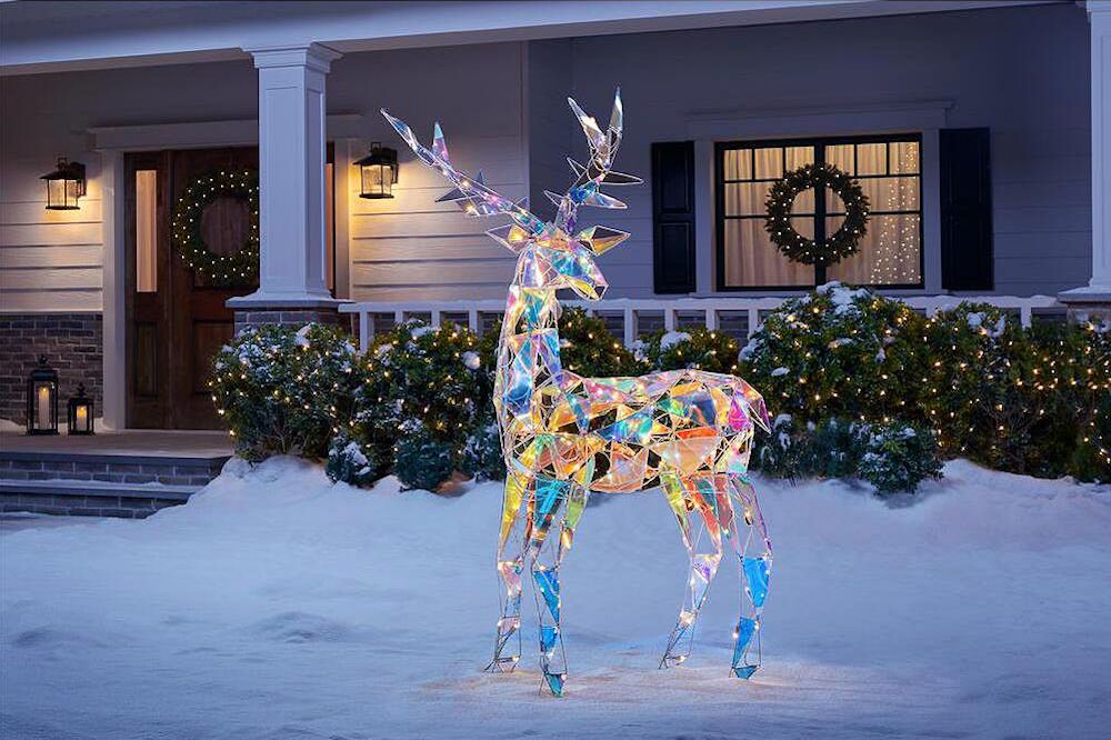72-inch Christmas sparkle modern angular-iridescent-reindeer buck standing in a snowy front yard in front of a classic white craftsman house with lights in the hedge and wreaths on the front door and window