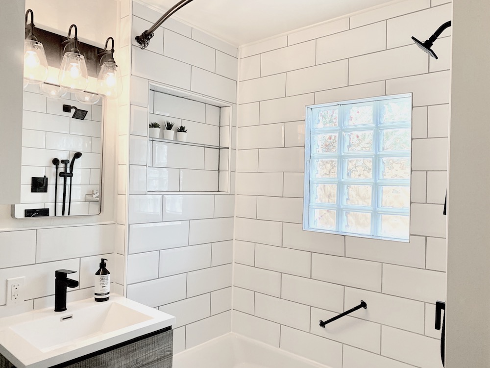 Beautiful modern bathroom with large white shower tile, white sink with a black faucet, grey edged vanity mirror, black vanity light and a recessed shelf with three small faux succulents