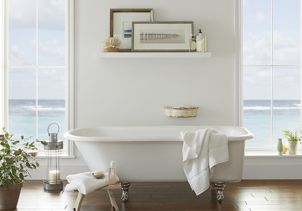 Beautiful traditional bathroom with large clawfoot bathtub, white display shelf with two frames and piece of coral, wood floors, two big windows looking out to the sea, and walls painted in BEHR White 52