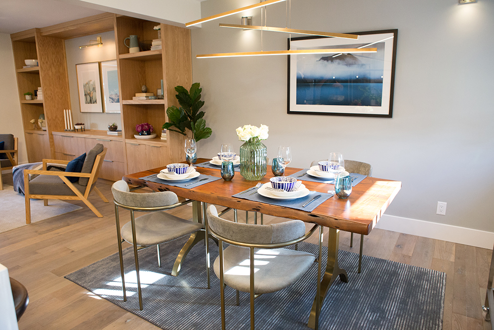 Chic dining room featured on The Property Brothers on HGTV with large wooden live edge dining table set with blue and white plates and bowls, four modern chairs, and modern abstract light fixture, a blue rug, a black framed photograph, and a set of built-in shelves