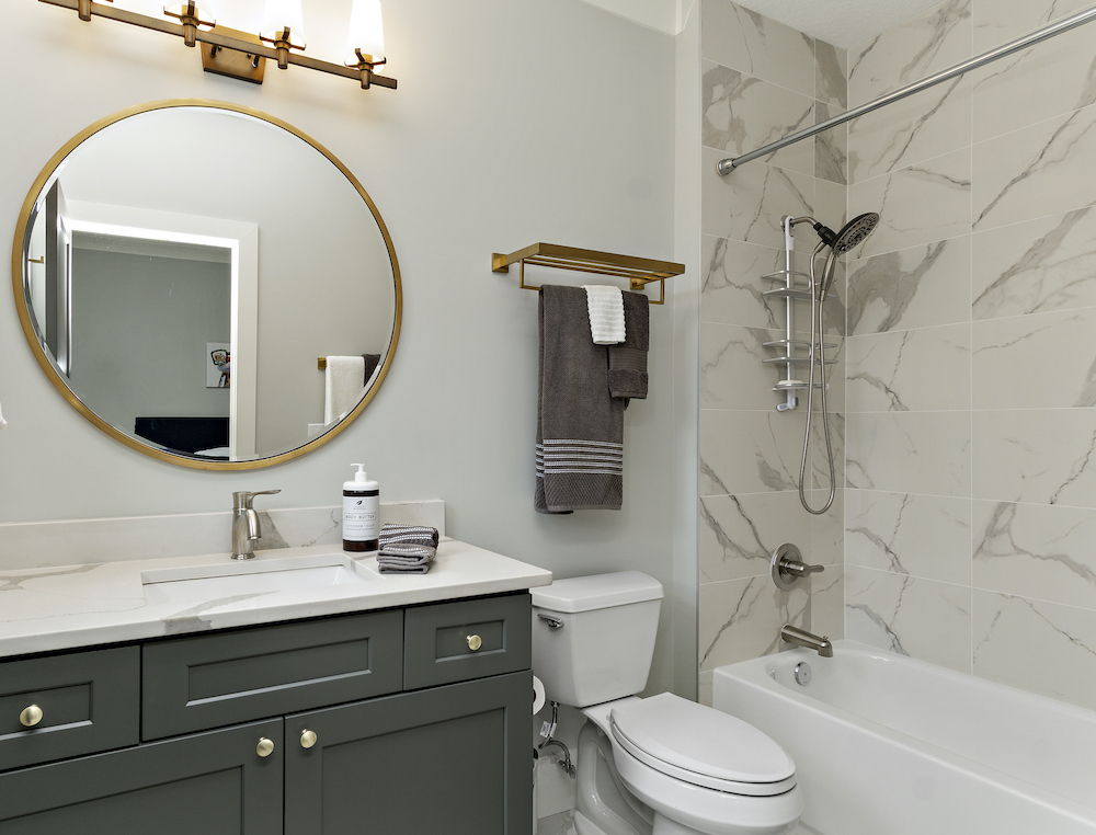 Simple chic bathroom with a dark grey vanity, gold circular wall mirror, a white toilet and a white bathtub shower combo with marbled wall tile