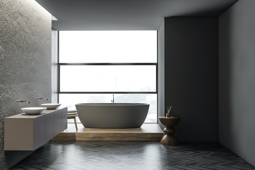 Interior of panoramic bathroom with concrete and gray walls, wooden floor, a wooden stool, a double vessel sinks standing on a grey floating vanity, and comfortable oval shaped standalone grey bathtub on a raised wooden platform in front of a wall of windows