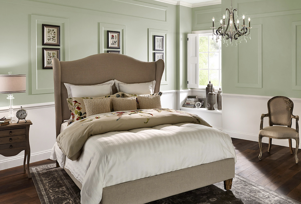 A charming bedroom with a large sand coloured upholstered bed, ornate black and crystal chandelier, six botanical drawings, and a walls painted in BEHR Polar Bear 75 and Sounds Of Nature S390-1