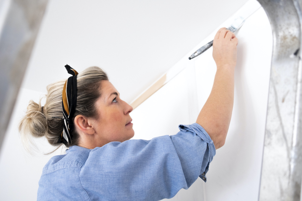 A woman wearing casual clothing and a head tie in her living room painting her wood panelled wall with white primer paint while standing on a ladder