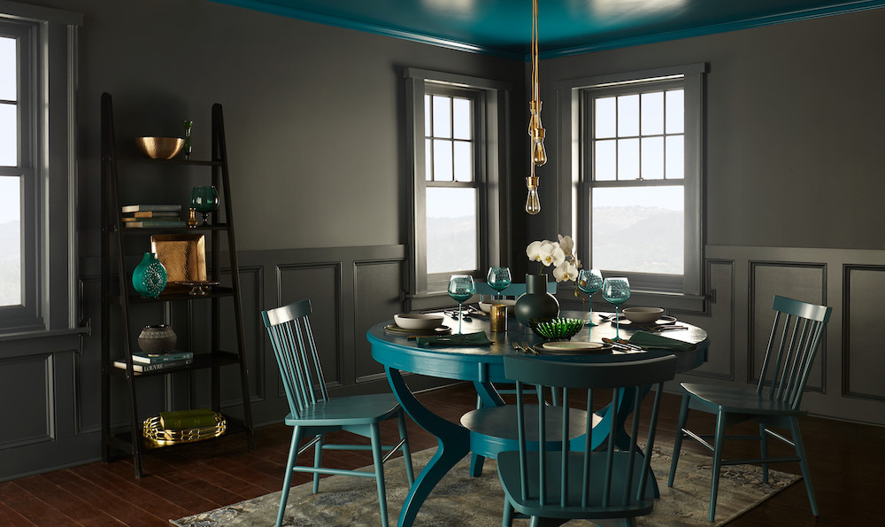 Chic dining room with a round blue table surrounded by four green chairs, two bright windows, a ladder shelf stacked with books and vessels, and walls painted in BEHR’s Blue Metal HDC-AC-25 interior paint colour