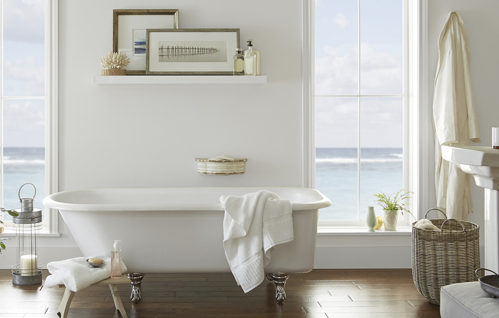 Charming bathroom with a large white clawfoot bathtub, two tall windows looking out to the sea, a small shelf with two frames, and walls painted in BEHR Seagull Gray N360-1 and White 52