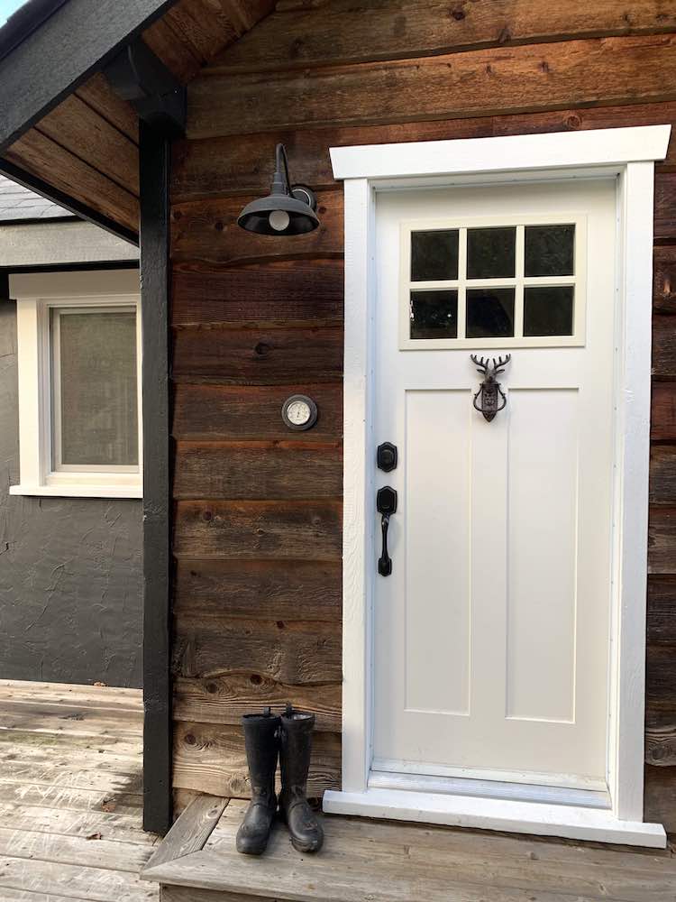 Exterior of a cottage with a bench with two cushions, a chair, a glass-topped table, and walls painted black with BEHR Black Smoke PPH-34 and window painted with BEHR Ultra Pure White 1850