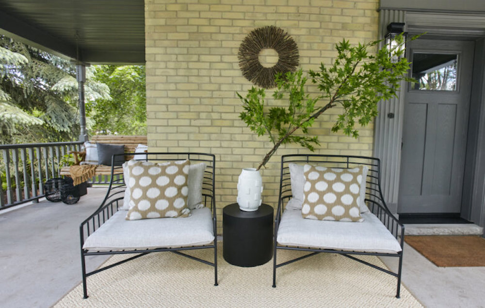 Beautiful conversation nook with two black metal chairs, cream and tan cushions, a white vase with a large green branch sitting on a black side table, on the front porch of a heritage home that was featured on HGTV’s Home to Win TV program