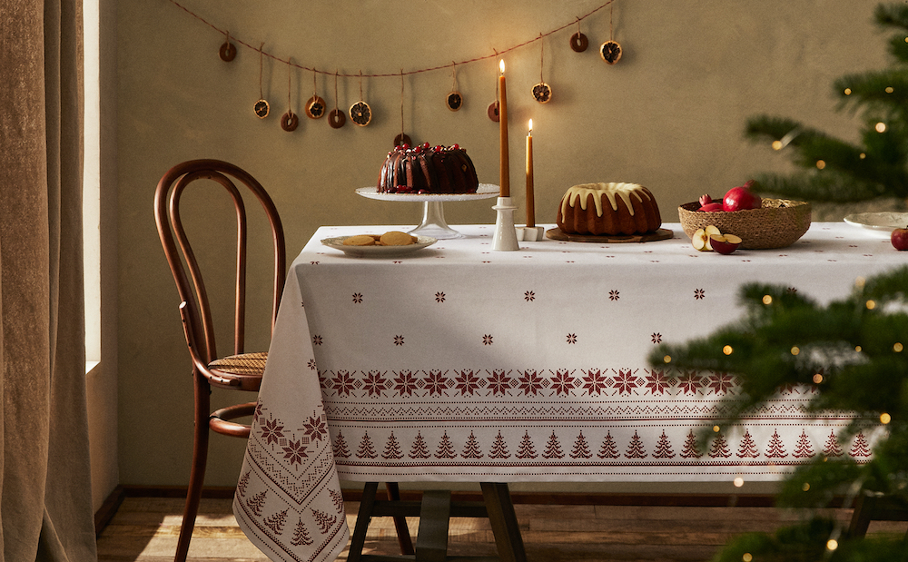 Christmas tablecloth with a contrast winter snowflake print with geometric shapes and fir trees onto of a table laid with Christmas cakes and baking, a wooden chair, two lit candles and a orange slice garland hanging on the wall behind