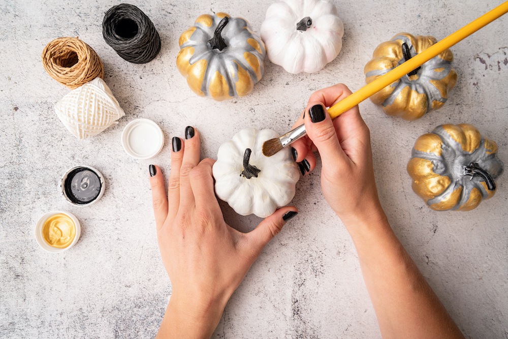 Woman does a Halloween DIY craft with a paintbrush colouring small pumpkins in white, gold and silver for a holiday party
