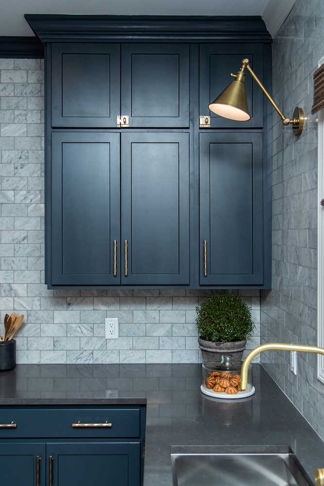 Beautiful kitchen with dark grey countertops, navy blue kitchen cabinets, a plate of macaroons, grey stone tile backsplash, and two gold wall scones