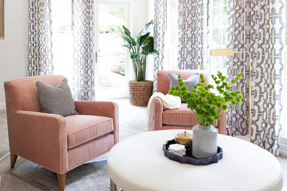 A light and airy living room with two pink velvet armchairs with grey throw pillow and white blanket, a round white ottoman with a tray and vase full of green flowers, patterned black and white curtains, and large palm house plant in the corner