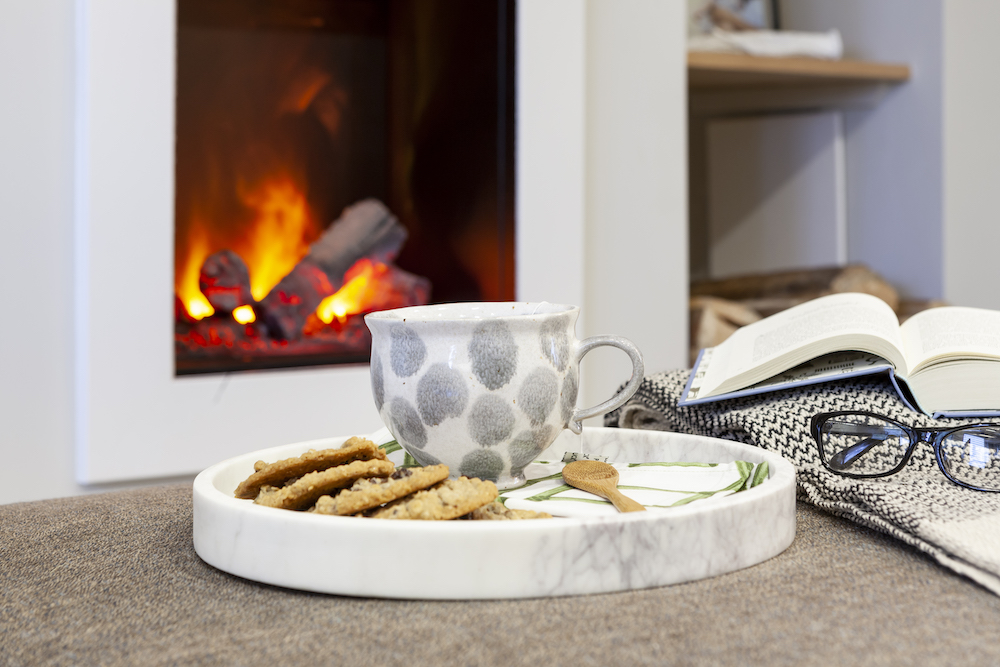 A pretty grey and white teacup sits on a marble tray with cookies in front of an electric fireplace