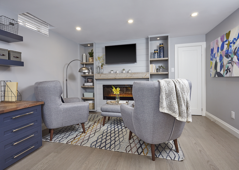Finished basement with chic at-home office and a lounge area with grey furnishings and large TV above an electric fireplace