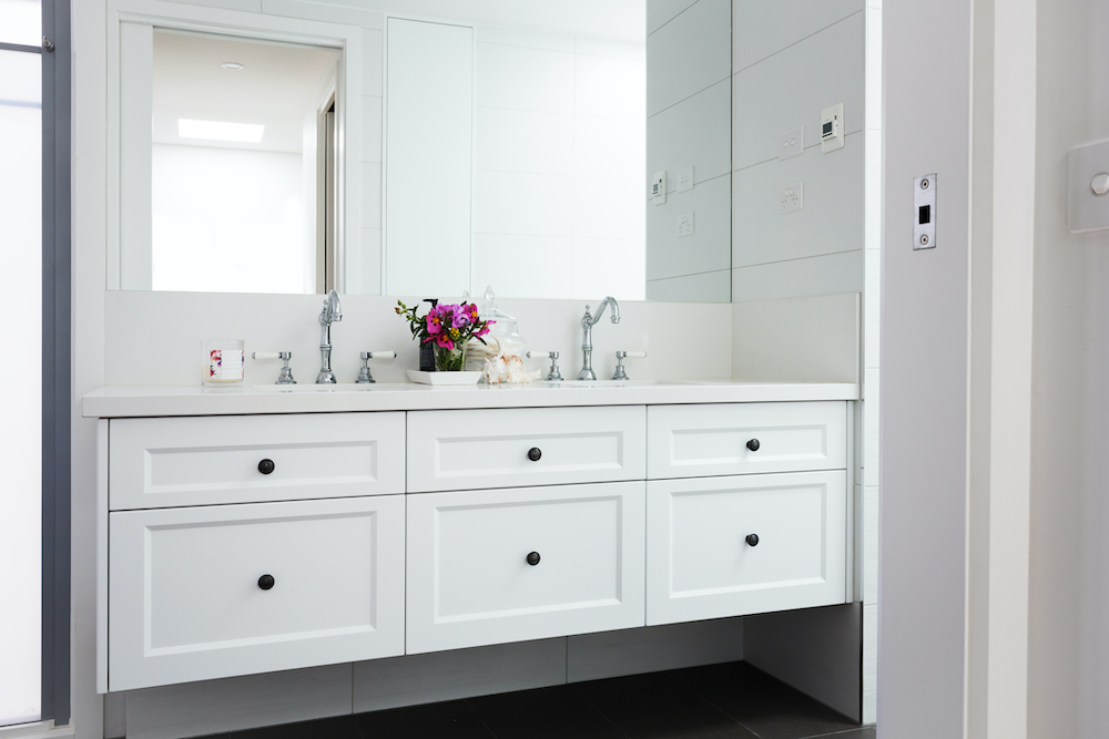 Beautiful white bathroom with large white floating vanity with quartz counter and six drawers, two sinks with silver coloured hardware, a vase of pink flowers, and a large wall mirror about it