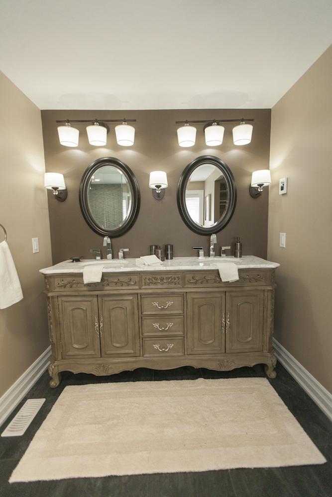 An ornate double sink vanity sits in a master ensuite bathroom with two oval mirrors and lots of lighting above it