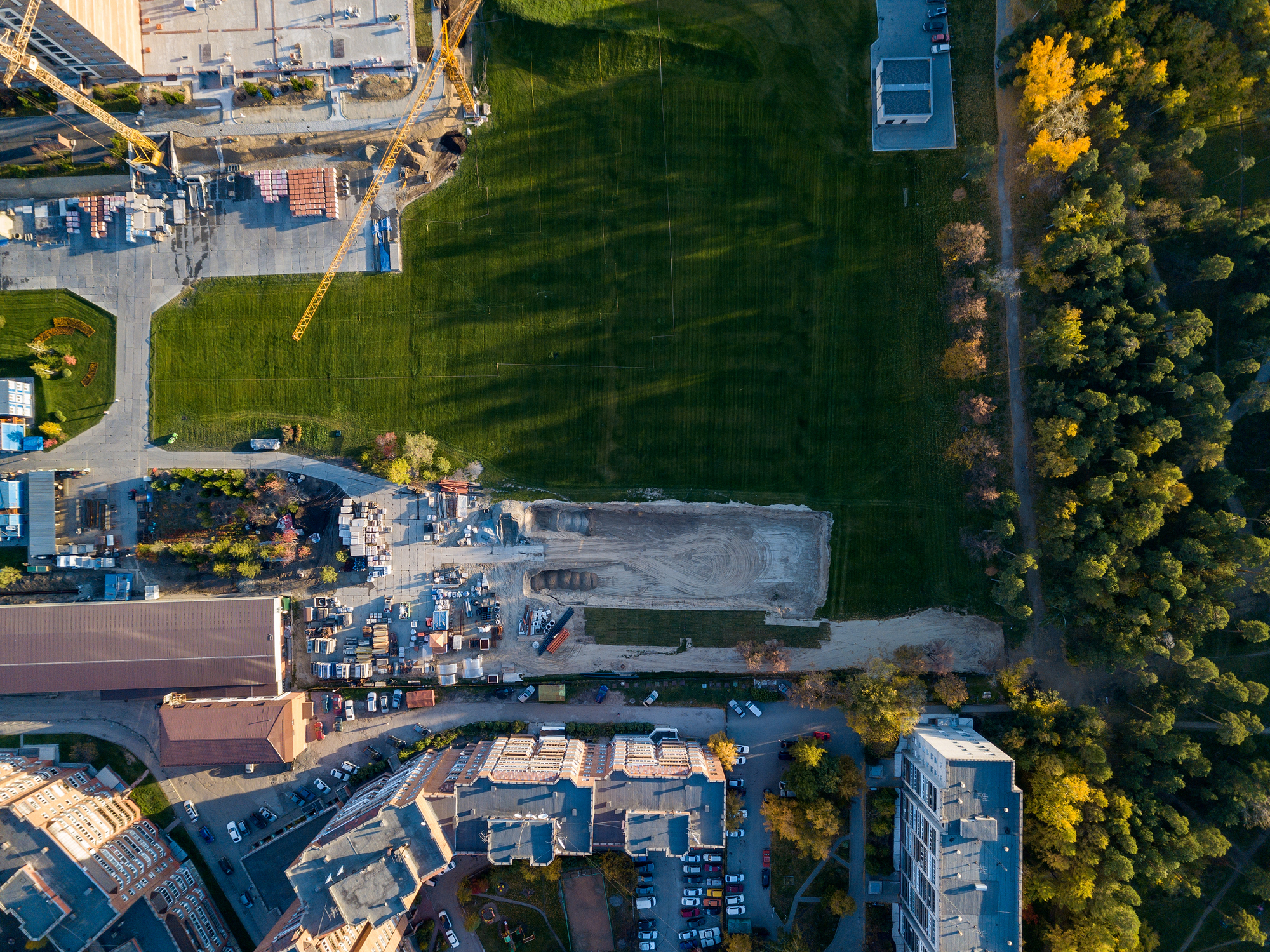 Aerial view of a building construction site at the storage of building materials surrounded by a green lawn with grass at sunset on an autumn sunny day on the border with forest and green trees