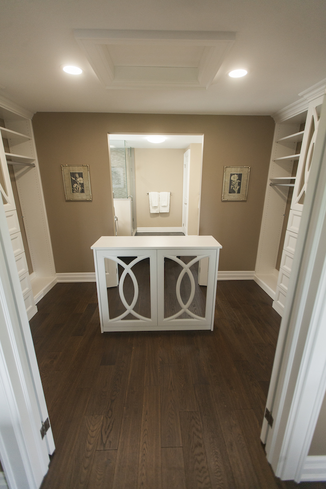 Large white walk in closet with dark wood floors, an ornate mirrored centre vanity and his and her hanging rails and drawers