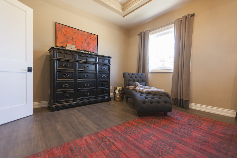 Master bedroom with a tall black dresser, flower painting, red carpet and grey tufted lounge seat