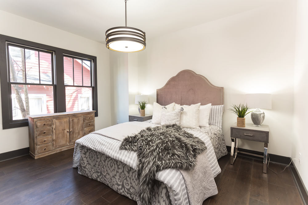 Masters of Flip luxe lodge bedroom bed with pink headboard, two grey bedside tables and a wooden dresser under large windows