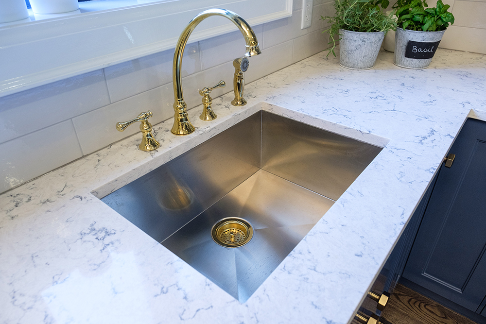 A deep, stainless-steel chef's sink with gold hardware and white quartz counterops