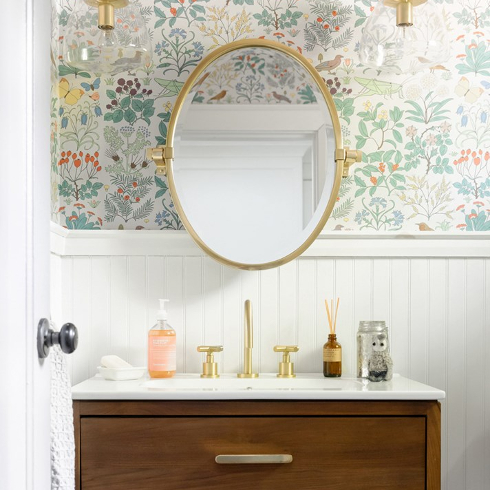 A powder room with wallpaper and a gold oval mirror
