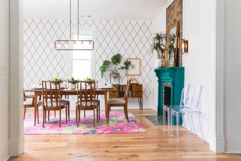 Black hanging light with exposed light bulbs in white dining room with wooden table topped with green plants standing on a large green and pink rug