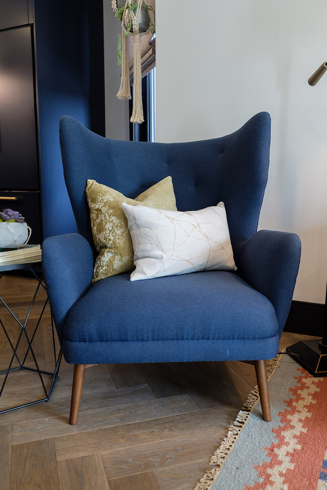 A chic mid century modern blue armchair with two throw pillows sits on a herringbone patter wood floor