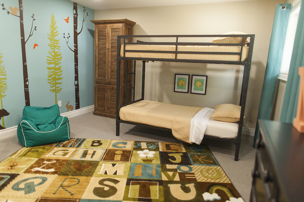 Cute kids room with bunk bed, teal coloured bean bag chair, wood dresser, alphabet rug and a fun woodsy decal mural on the wall