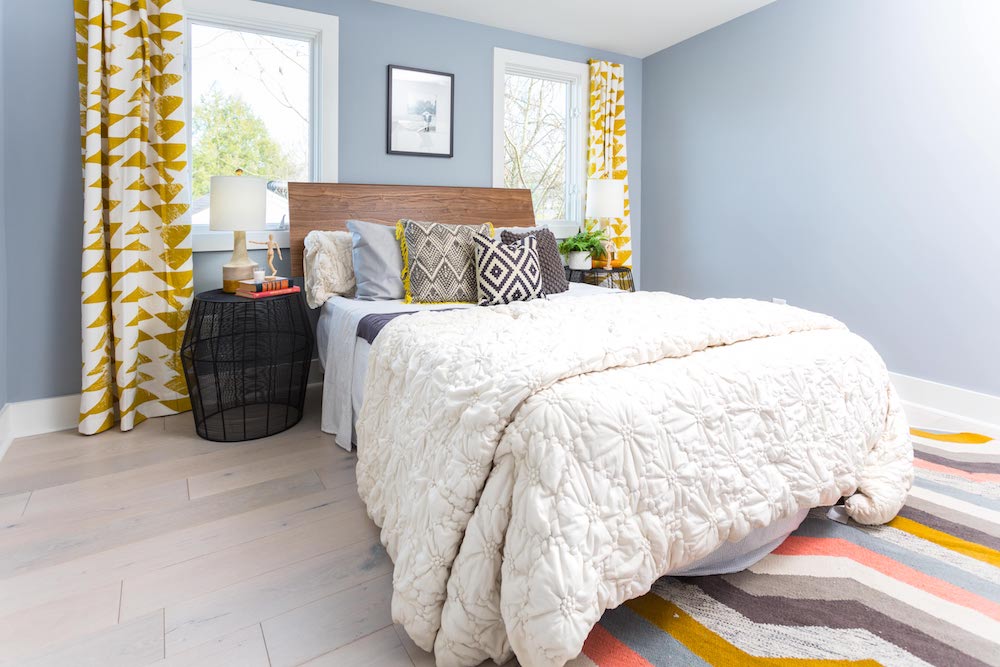 Masters of Flip train station house bedroom with yellow patterned curtains, a bed with a wooden headboard and grey-blue walls